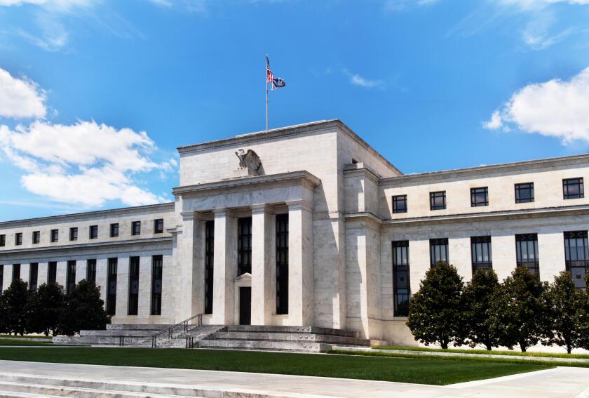 Exterior view of Federal Reserve Board of Governors Building facade entrance with eagle stone statue and American flags over a blue cloudscape sky.