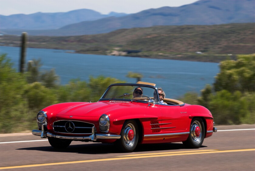 A 1958 Mercedes Benz 300SL Roadster driving along a scenic highway near a lake.