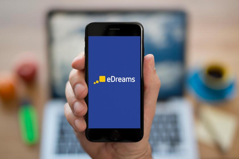 A man looks at his iPhone which displays the eDreams logo, while sat at his computer desk (Editorial use only).