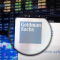 Goldman Sachs company logo on a website with blurry stock market developments in the background, seen on a computer screen through a magnifying glass