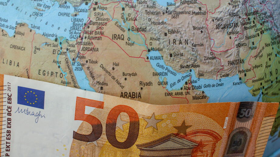 A fifty euro bill placed over the Strait of Hormuz on a world globe, symbolizing the impact of Middle East geopolitical tensions on European fuel pric